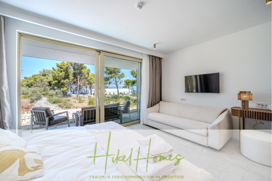 Hochwertige Meerblick Villa SUN in Sutivan (Insel Brac) – 3 SZ + Bad, Pool, Sommerküche, WC, … - Ein helles, modernes Schlafzimmer mit einem weißen Bett und einem Sofa, einem an der Wand montierten Fernseher und großen Glastüren, die sich zu einer Terrasse mit Sitzgelegenheiten im Freien öffnen, mit Blick auf üppige Bäume und das Meer. Das Logo "Hikel Homes" befindet sich am Boden. (44996)