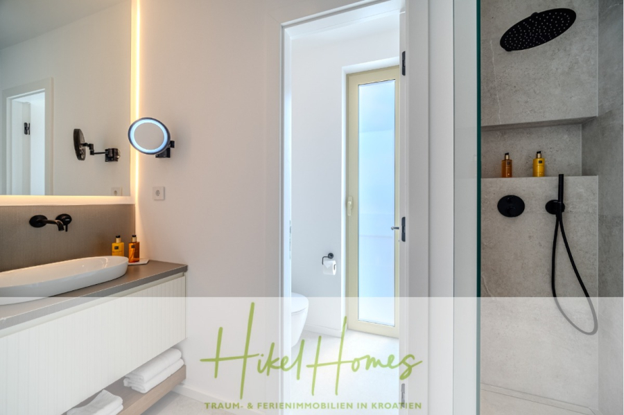 Hochwertige Meerblick Villa SUN in Sutivan (Insel Brac) – 3 SZ + Bad, Pool, Sommerküche, WC, … - Modernes Badezimmer mit einem Waschbecken, einem runden Spiegel, einer Toilette, einer Dusche mit schwarzen Armaturen und Toilettenartikeln; weiße Wände und sauberes Design. Der Text "Hikel Homes" und ein deutscher Slogan erscheinen am unteren Rand. (44999)