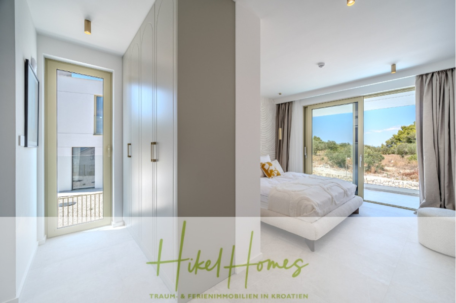 Hochwertige Meerblick Villa SUN in Sutivan (Insel Brac) – 3 SZ + Bad, Pool, Sommerküche, WC, … - Helles, modernes Schlafzimmer mit großen Fenstern, weißen Möbeln, Einbauschränken und Blick auf Bäume und Felsen draußen. Der Text auf dem Bild lautet "Hikel Homes, Traum & Ferienimmobilien in Kroatien". (44992)