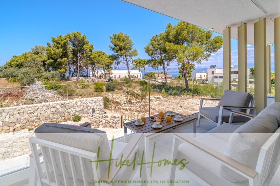Hochwertige Meerblick Villa SUN in Sutivan (Insel Brac) – 3 SZ + Bad, Pool, Sommerküche, WC, … - Eine moderne Terrasse mit weißen Gartenmöbeln, einem für das Frühstück gedeckten Tisch und einem Blick auf Bäume, Steinmauern und Häuser unter einem strahlend blauen Himmel. Der Text "Hikel Homes" erscheint am unteren Rand. (44995)