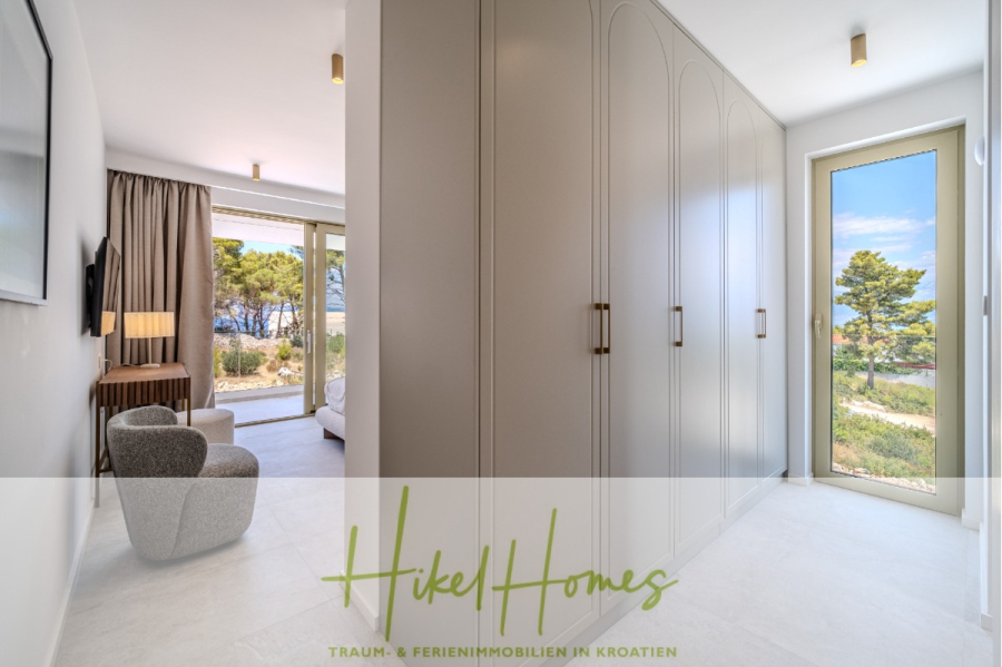 Hochwertige Meerblick Villa SUN in Sutivan (Insel Brac) – 3 SZ + Bad, Pool, Sommerküche, WC, … - Modernes, helles Schlafzimmer mit weißen Schränken, einem gemütlichen Sessel, großen Fenstern und einem Blick auf Bäume und das Meer draußen. Logo und Text von "Hikel Homes" erscheinen am unteren Rand des Bildes. (44997)