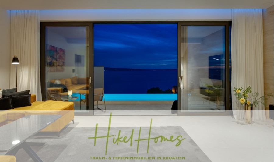 Sofort einziehen! Meerblick Villa CECILIE Infinity Pool, 3 SZ, Sommerküche, Videoraum mit PS5, …. - Modernes Wohnzimmer mit großen Glastüren, die auf eine Terrasse, einen Infinity-Pool und einen malerischen Meerblick in der Abenddämmerung führen. Der Innenraum bietet gemütliche Sitzgelegenheiten und modernes Dekor. Unten steht „Hikel Homes“. (43921)