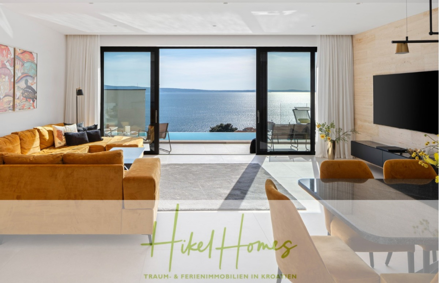 Sofort einziehen! Meerblick Villa CECILIE Infinity Pool, 3 SZ, Sommerküche, Videoraum mit PS5, …. - Modernes Wohnzimmer mit senfgelben Sofas, großen Fenstern mit Blick auf das blaue Meer, einem an der Wand montierten Fernseher, zeitgenössischer Einrichtung und hereinströmendem Sonnenlicht. Das Logo unten lautet „Hikel Homes – Traum- & Ferienimmobilien in Kroatien“. (43900)