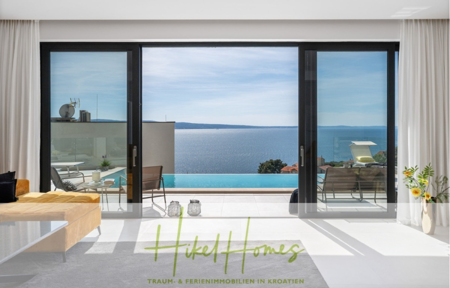Sofort einziehen! Meerblick Villa CECILIE Infinity Pool, 3 SZ, Sommerküche, Videoraum mit PS5, …. - Modernes Wohnzimmer mit großen Glastüren, die auf eine Terrasse, einen Infinity-Pool und einen malerischen Meerblick unter klarem Himmel führen. Unten steht der Text: „Hikel Homes Traum- & Ferienimmobilien in Kroatien.“. (43894)