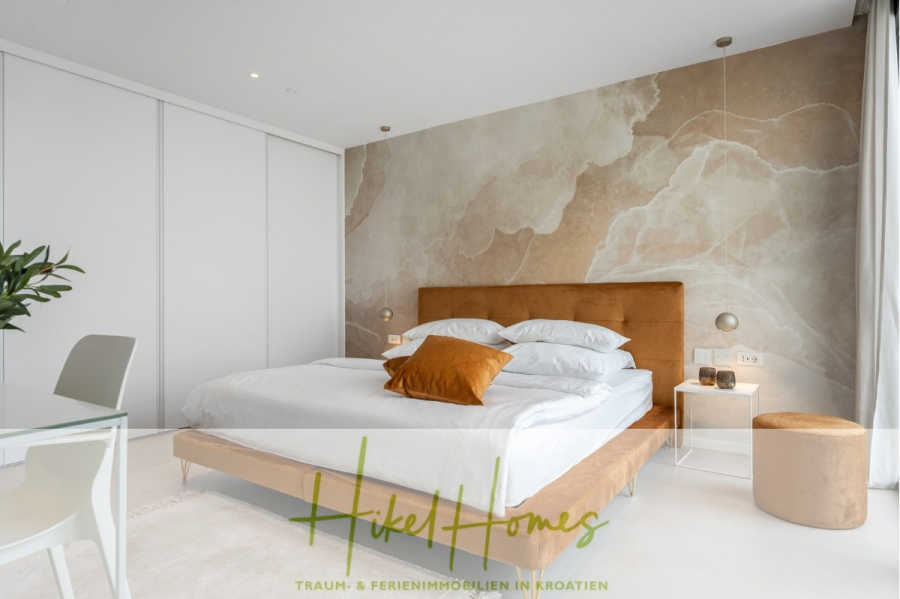 Sofort einziehen! Meerblick Villa CECILIE Infinity Pool, 3 SZ, Sommerküche, Videoraum mit PS5, …. - Modernes Schlafzimmer mit beigefarbenem Polsterbett, weißer Bettwäsche, grünem Akzentkissen, abstraktem Wandbild, Schiebetüren, weißem Schreibtisch und Stuhl, Topfpflanze und großen Fenstern mit transparenten Vorhängen. Unten das Logo „Hikel Homes“. (43911)
