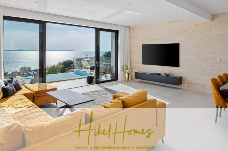 Sofort einziehen! Meerblick Villa CECILIE Infinity Pool, 3 SZ, Sommerküche, Videoraum mit PS5, …. - Modernes Wohnzimmer mit gelbem Ecksofa, Fernseher und großen Glastüren, die auf eine Terrasse mit atemberaubendem Meerblick führen. Sonnenlicht durchflutet den Raum, und ein Essbereich mit senffarbenen Stühlen ist sichtbar. Der Text „Hikel Homes“ ist eingeblendet. (43897)