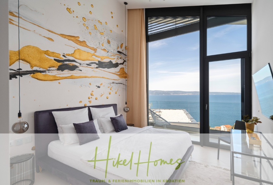 Sofort einziehen! Meerblick Villa CECILIE Infinity Pool, 3 SZ, Sommerküche, Videoraum mit PS5, …. - Modernes Schlafzimmer mit großem Bett, abstrakter Wandkunst in Gold und Schwarz, raumhohen Fenstern und Blick aufs Meer. Am unteren Bildrand sind der deutsche Schriftzug und Slogan „Hikel Homes“ zu sehen. (43910)