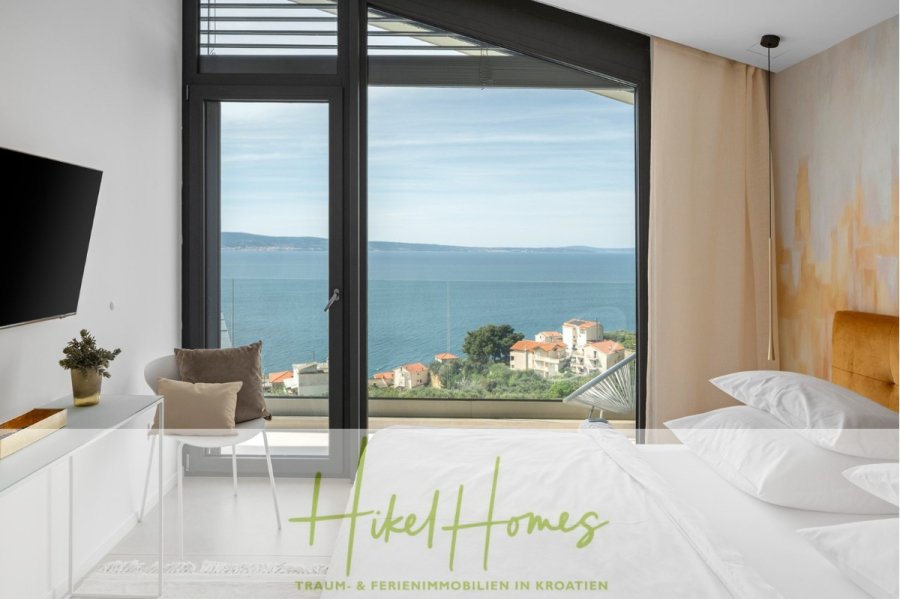 Sofort einziehen! Meerblick Villa CECILIE Infinity Pool, 3 SZ, Sommerküche, Videoraum mit PS5, …. - Modernes Schlafzimmer mit großen Fenstern und Blick auf das Meer, Küstengebäude und den blauen Himmel. Minimalistisches Dekor mit weißem Bett, orangefarbenem Sessel und Wandfernseher. Die Textüberlagerung lautet „Hikel Homes“ und „Traum- & Ferienimmobilien in Kroatien“. (43903)