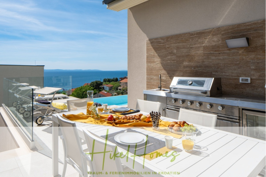 Sofort einziehen! Meerblick Villa CECILIE Infinity Pool, 3 SZ, Sommerküche, Videoraum mit PS5, …. - Ein moderner Essbereich im Freien mit gedecktem Tisch, Grill und Sonnenliegen bietet unter einem klaren, sonnigen Himmel Blick auf das Meer und die Küstenhäuser. Die Umgebung ist hell und einladend. (43896)