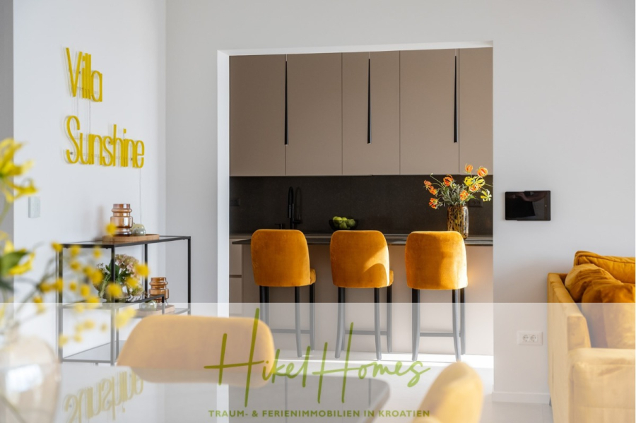 Sofort einziehen! Meerblick Villa CECILIE Infinity Pool, 3 SZ, Sommerküche, Videoraum mit PS5, …. - Moderne Küche mit beigen Schränken und drei gelben Barhockern. Auf der Theke steht eine Vase mit Blumen. „Villa Sunshine“ steht in gelben Buchstaben an der Wand, und Sonnenlicht erhellt das weiß-gelbe Dekor. (43901)