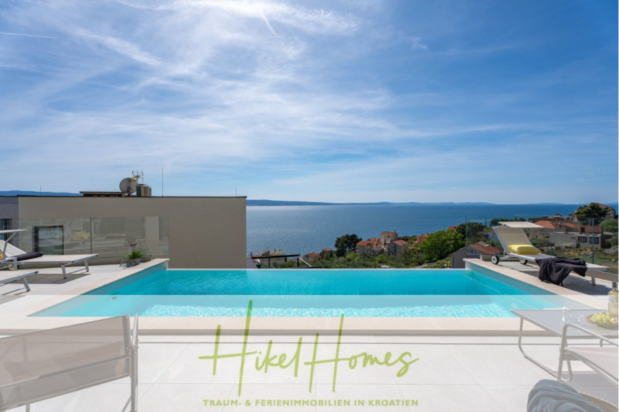 Sofort einziehen! Meerblick Villa CECILIE Infinity Pool, 3 SZ, Sommerküche, Videoraum mit PS5, …. - Moderner Pool auf dem Dach mit Liegestühlen und Blick auf das Meer und die Dächer der Küste unter einem klaren blauen Himmel. Auf dem Pooldeck steht in Grün und Gold „Hikel Homes – Traum- & Ferienimmobilien in Kroatien“. (43888)