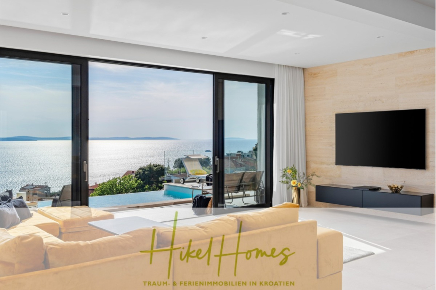 Sofort einziehen! Meerblick Villa CECILIE Infinity Pool, 3 SZ, Sommerküche, Videoraum mit PS5, …. - Modernes Wohnzimmer mit gelbem Sofa, großem Fernseher und raumhohen Glastüren, die auf eine Terrasse mit Pool, Liegestühlen und Panoramablick auf das Meer führen. „Hikel Homes“-Logo und deutscher Text unten. (43909)