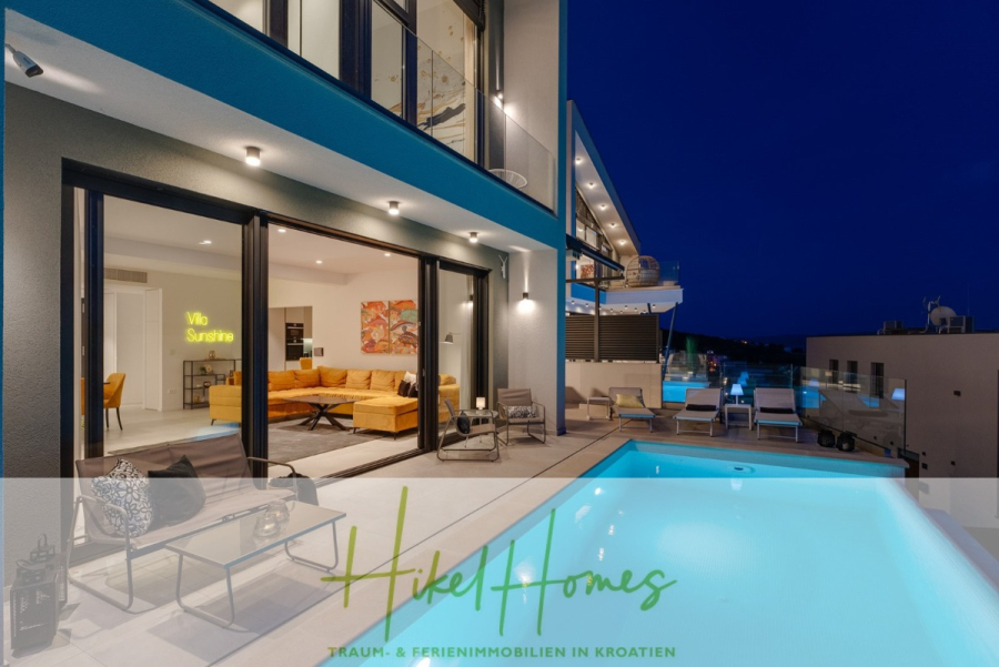 Sofort einziehen! Meerblick Villa CECILIE Infinity Pool, 3 SZ, Sommerküche, Videoraum mit PS5, …. - Moderne Villa bei Nacht mit beleuchtetem Pool, offenen Schiebetüren zum stilvollen Wohnzimmer, zeitgenössischen Möbeln und einem darüberliegenden Balkon. Unten erscheint der Text „Hikel Homes – Traum- & Ferienimmobilien in Kroatien“. (43922)