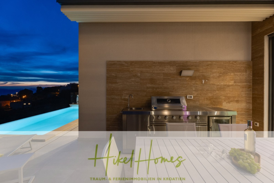 Sofort einziehen! Meerblick Villa CECILIE Infinity Pool, 3 SZ, Sommerküche, Videoraum mit PS5, …. - Moderne Außenküche mit Edelstahlgrill auf einer Steinterrasse, neben einem Swimmingpool mit Blick auf einen malerischen Sonnenuntergang. Das Logo „Hike Homes“ und deutscher Text erscheinen unten im Bild. (43908)