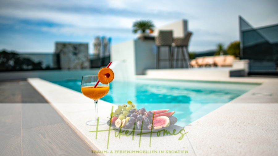 Luxus trifft Meerblick – Ein Anwesen der Extraklasse – Sofort einziehen - Ein Glas Orangensaft mit einer Fruchtgarnitur und ein Teller mit frischem Obst stehen an einem modernen Außenpool. Sonniges Wetter, Palmen und stilvolle Möbel sind zu sehen. Der Text "Hikel Homes Traum- & Ferienimmobilien in Kroatien" ist überlagert. (46474)