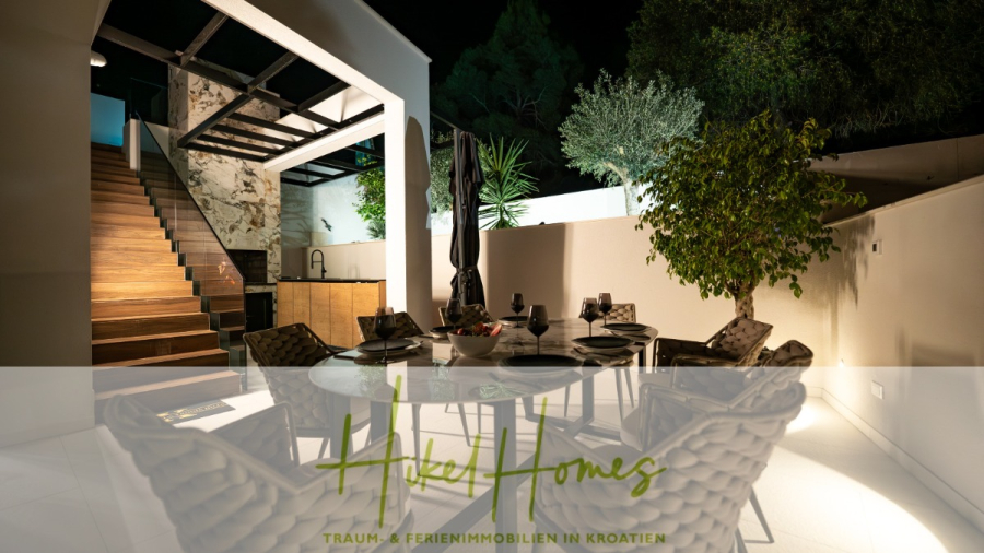 Luxus trifft Meerblick – Ein Anwesen der Extraklasse – Sofort einziehen - Eleganter Essbereich im Freien mit einem gedeckten Tisch unter einer Pergola, umgeben von üppigem Grün und stimmungsvoller Beleuchtung. Links führt eine Treppe nach oben, und unten steht der Text „Traum- & Ferienimmobilien in Kroatien“. (43751)