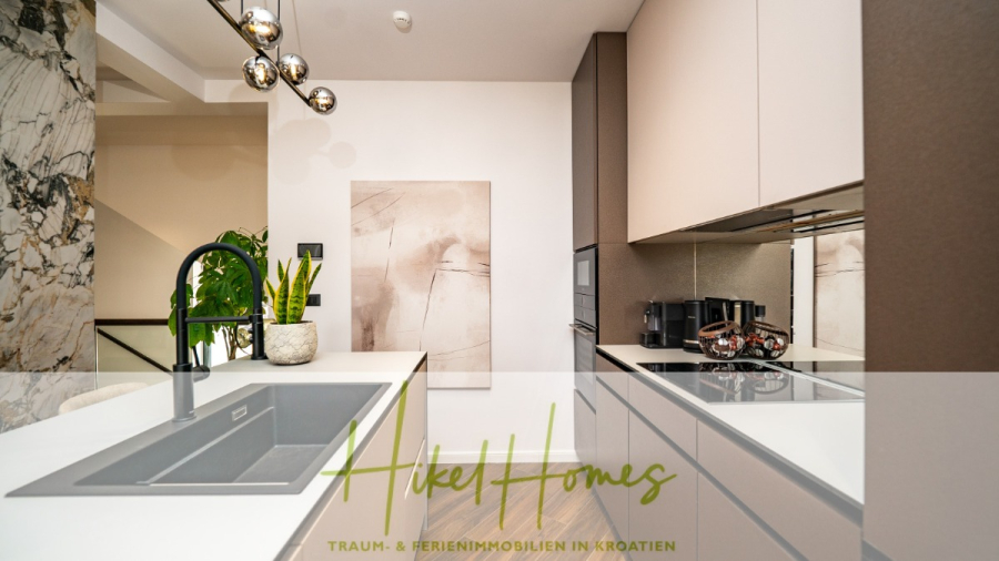 Luxus trifft Meerblick – Ein Anwesen der Extraklasse – Sofort einziehen - Moderne Küche mit beigen Schränken, schwarzem Wasserhahn, eingebauter Spüle, Kaffeemaschinen und Marmorakzenten. Abstrakte Kunst hängt an der Wand, eine Pflanze sorgt für Grünzeug. Der Text "Aikel Homes - Traum- & Ferienimmobilien in Kroatien" überlagert das Bild. (46497)