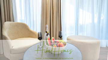 Eine gemütliche Sitzecke mit zwei beigen Stühlen und einem kleinen Tisch mit einer Obstschale, zwei Weingläsern und einer Flasche Sekt vor hellen Vorhängen. Der Text auf dem Bild lautet: "Hikel Homes - Traum- & Ferienimmobilien in Kroatien. - Luxus trifft Meerblick – Ein Anwesen der Extraklasse – Sofort einziehen