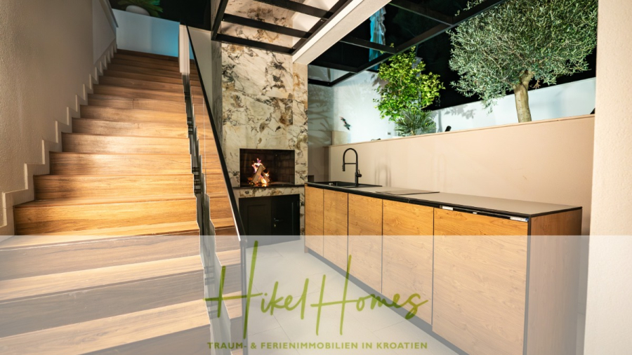 Luxus trifft Meerblick – Ein Anwesen der Extraklasse – Sofort einziehen - Moderne Küchenzeile im Innenbereich mit Holzschränken, schwarzer Arbeitsplatte und einem eleganten Waschbecken neben einer Treppe; Marmor-Akzentwand und große Fenster mit Grünpflanzen im Außenbereich. Das Logo "Hikel Homes" überlagert das Bild. (46500)