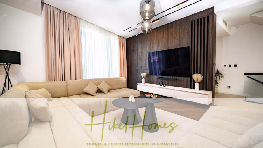 Luxus trifft Meerblick – Ein Anwesen der Extraklasse – Sofort einziehen - Modernes Wohnzimmer mit einem großen beigefarbenen Sofa, einem runden grauen Couchtisch und einem Flachbildfernseher an einer Holzwand. Von der Decke hängen Lampen, und am Fenster hängen hohe rosa Vorhänge. (43741)