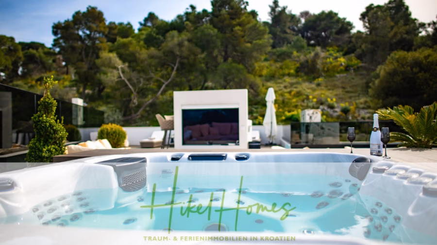 Luxus trifft Meerblick – Ein Anwesen der Extraklasse – Sofort einziehen - Whirlpool mit sprudelndem Wasser auf einer Holzterrasse, umgeben von üppigem Grün und Gartenmöbeln. Im Hintergrund ein Flachbildfernseher und eine Champagnerflasche. Darüber sonniger, klarer Himmel. Unten das Logo „Hikel Homes“. (43738)