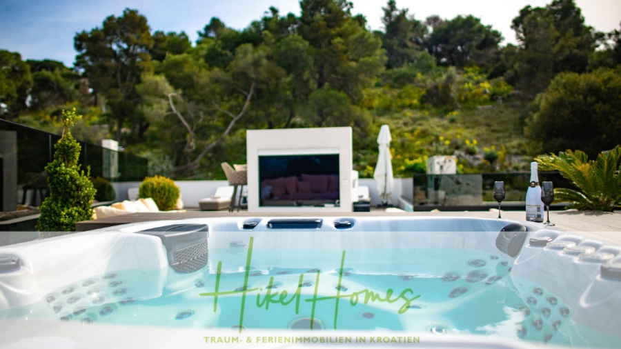 Luxus trifft Meerblick – Ein Anwesen der Extraklasse – Sofort einziehen - Luxuriöser Whirlpool im Freien mit sprudelndem Wasser auf einer sonnigen Terrasse; Sektflasche und Gläser an der Seite, mit modernen Sitzgelegenheiten im Freien, Fernseher und üppigem grünen Wald im Hintergrund. Textüberlagerung "Hikel Homes" auf der Vorderseite. (46489)