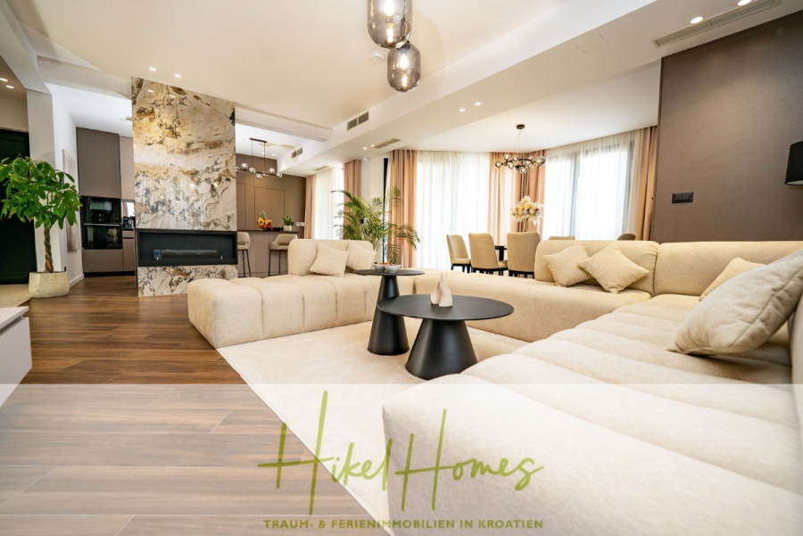 Luxus trifft Meerblick – Ein Anwesen der Extraklasse – Sofort einziehen - Modernes, geräumiges Wohnzimmer mit einem großen beigen Sektionssofa, zwei schwarzen runden Couchtischen, Holzfußboden und großen Fenstern mit transparenten Vorhängen; Essbereich und Küche sind im Hintergrund zu sehen. (46494)