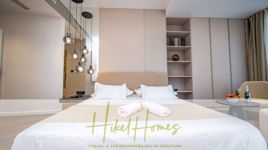 Luxus trifft Meerblick – Ein Anwesen der Extraklasse – Sofort einziehen - Ein modernes Schlafzimmer mit einem sauber gemachten Doppelbett, zwei gerollten Handtüchern und Hängelampen. Das Zimmer ist in neutralen Farben gehalten und verfügt über eingebaute Regale und einen kleinen Beistelltisch mit einer Obstschale und einem gekühlten Getränk. Der Text lautet „Hikel Homes“. (43727)