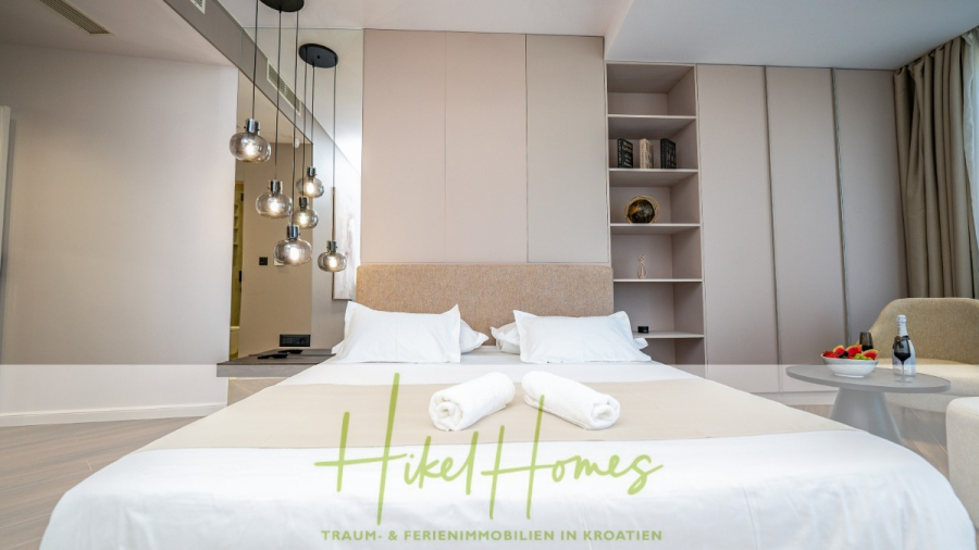 Luxus trifft Meerblick – Ein Anwesen der Extraklasse – Sofort einziehen - Ein modernes Hotelzimmer verfügt über ein ordentlich gemachtes Doppelbett mit zwei gerollten Handtüchern, Pendelleuchten, offene Regale und einen Sitzbereich mit Champagner und Erdbeeren. Der Schriftzug "Hikel Homes" ist am unteren Rand eingeblendet. (46477)