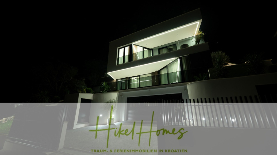 Luxus trifft Meerblick – Ein Anwesen der Extraklasse – Sofort einziehen - Modernes Haus mit geometrischer Architektur, das bei Nacht beleuchtet wird. Davor sind ein Zaun und ein angelegter Garten zu sehen. Der überlagerte Text lautet "Hikel Homes - Traum- & Ferienimmobilien in Kroatien". (46502)