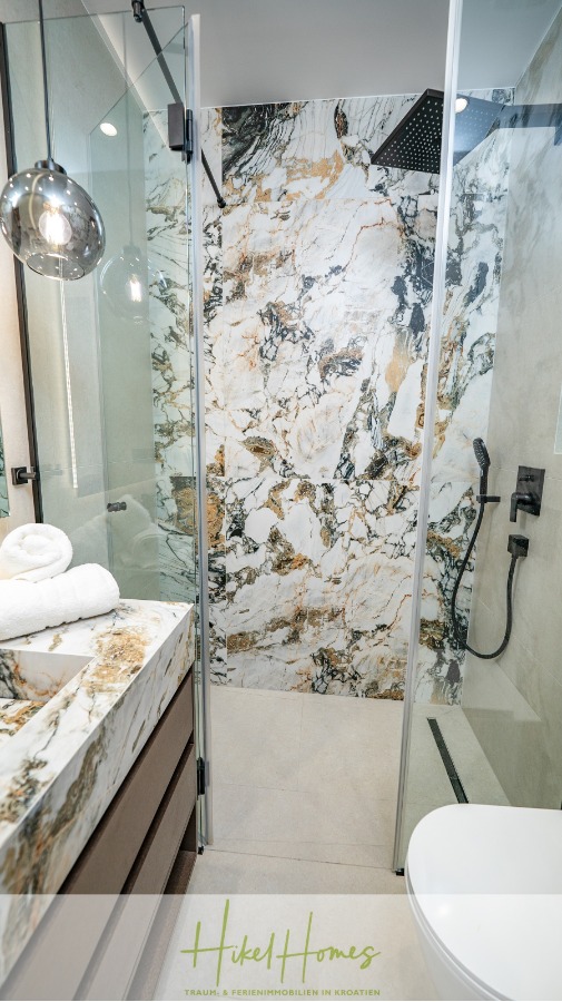 Luxus trifft Meerblick – Ein Anwesen der Extraklasse – Sofort einziehen - Ein modernes Badezimmer mit einer Glasdusche, markanten Marmorwänden und -waschbecken, einer schwarzen Duscharmatur, einer Hängelampe, gefalteten weißen Handtüchern und neutralem Bodenbelag. (46481)