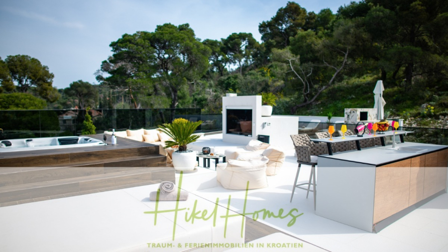 Luxus trifft Meerblick – Ein Anwesen der Extraklasse – Sofort einziehen - Eine moderne Außenterrasse mit weißen Liegestühlen, einem Whirlpool, einem Essbereich und einem Grill, umgeben von üppig grünen Bäumen unter einem klaren Himmel. Der Text auf dem Bild lautet: "Hikel Homes - Traum- & Ferienimmobilien in Kroatien. (46488)