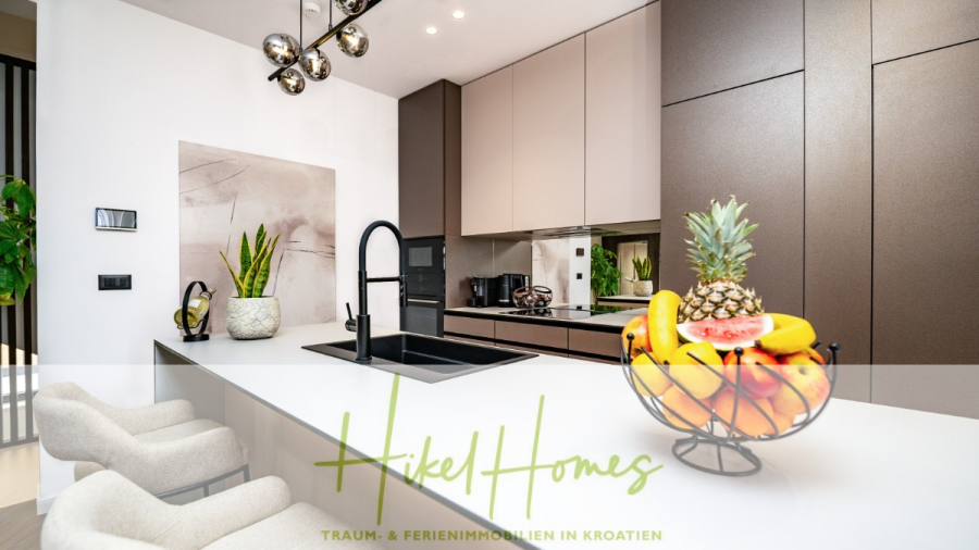 Luxus trifft Meerblick – Ein Anwesen der Extraklasse – Sofort einziehen - Moderne Küche mit neutralen Schränken, einer weißen Arbeitsplatte auf einer Insel mit schwarzem Waschbecken, zwei beigen Stühlen, einer dekorativen Obstschale und Zimmerpflanzen. Das Bild enthält einen Text: "Hikel Homes Traum- & Ferienimmobilien in Kroatien. (46496)