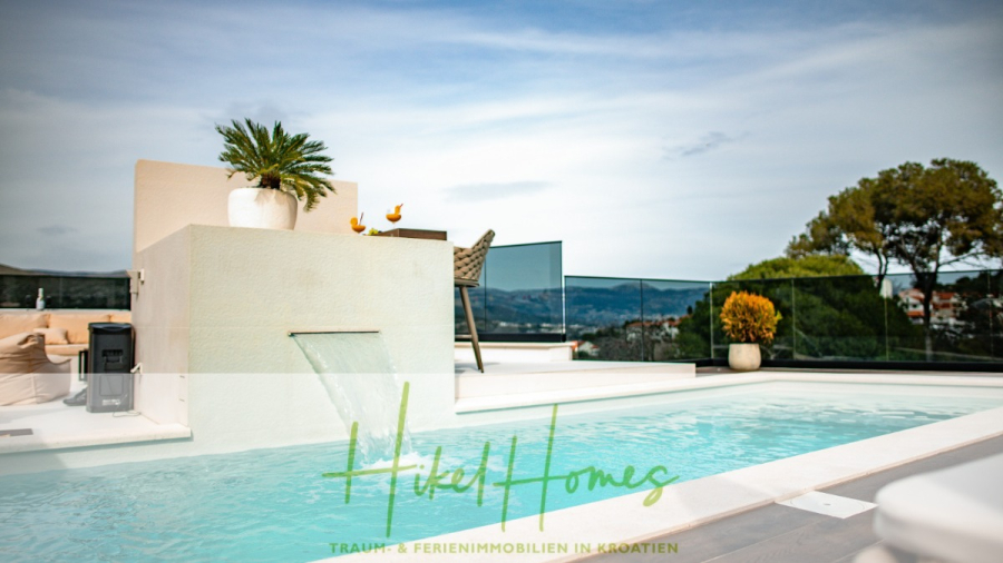 Luxus trifft Meerblick – Ein Anwesen der Extraklasse – Sofort einziehen - Ein modernes Freibad mit einem kleinen Wasserfall, Topfpflanzen und einem durchsichtigen Glaszaun mit Blick auf Bäume und eine entfernte Stadt, unter einem teilweise bewölkten Himmel. Der Text am unteren Rand lautet "Hotel Homes". (46490)