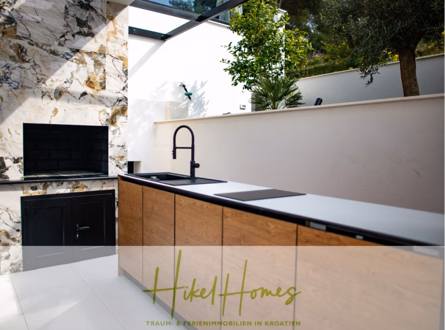 Luxus trifft Meerblick – Ein Anwesen der Extraklasse – Sofort einziehen - Moderne Außenküche mit Marmorrückwand, eleganten Holzschränken und schwarzem Wasserhahn. Ein eingebauter Grill ist vorhanden und der Bereich ist von Grün umgeben. Der Raum ist offen und gut beleuchtet. (43736)