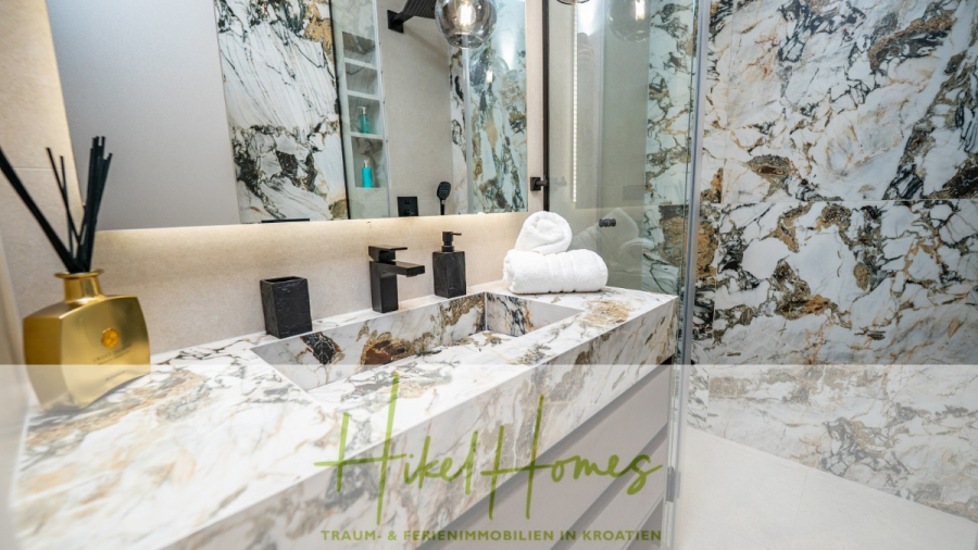 Luxus trifft Meerblick – Ein Anwesen der Extraklasse – Sofort einziehen - Luxuriöses, modernes Badezimmer mit Marmorarbeitsflächen und -wänden, einem Doppelwaschbecken, schwarzen Armaturen, einer Glasdusche und aufgerollten weißen Handtüchern auf dem Tresen. Dekorative Elemente und sanfte Beleuchtung sorgen für Eleganz. (46480)