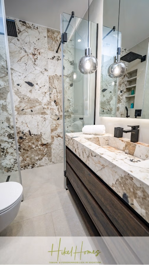 Luxus trifft Meerblick – Ein Anwesen der Extraklasse – Sofort einziehen - Modernes Badezimmer mit Steinmuster an Waschtisch und Wand sowie einer Duschtür aus Glas. Zwei Hängeleuchten erhellen den Raum. Eine weiße Toilette und ordentlich gefaltete Handtücher unterstreichen das klare, elegante Design. (43733)
