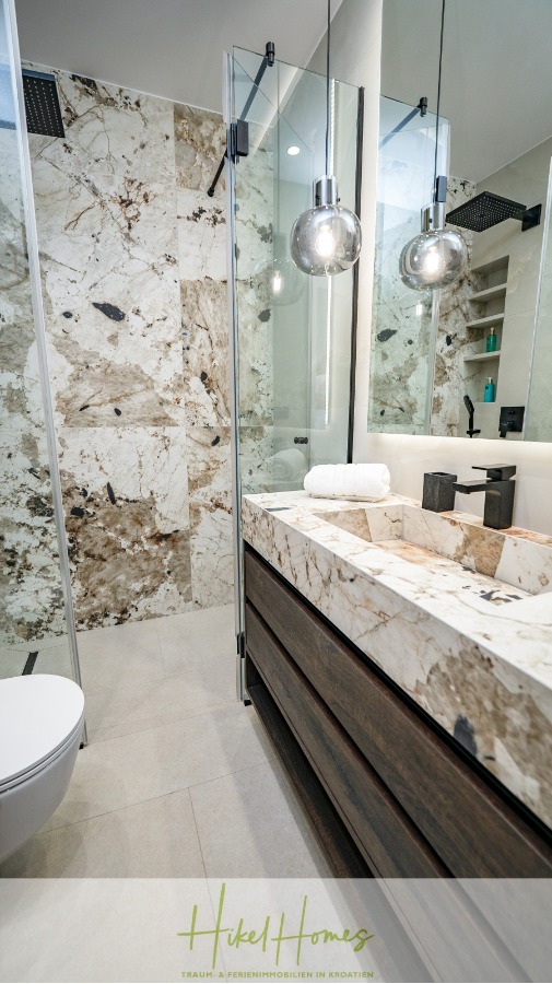 Luxus trifft Meerblick – Ein Anwesen der Extraklasse – Sofort einziehen - Modernes Badezimmer mit Marmorwänden und -arbeitsplatte, Glasduschabtrennung, runden Hängelampen, Holzschubladen, weißer Toilette und ordentlich gefalteten Handtüchern auf dem Waschbecken. (46484)