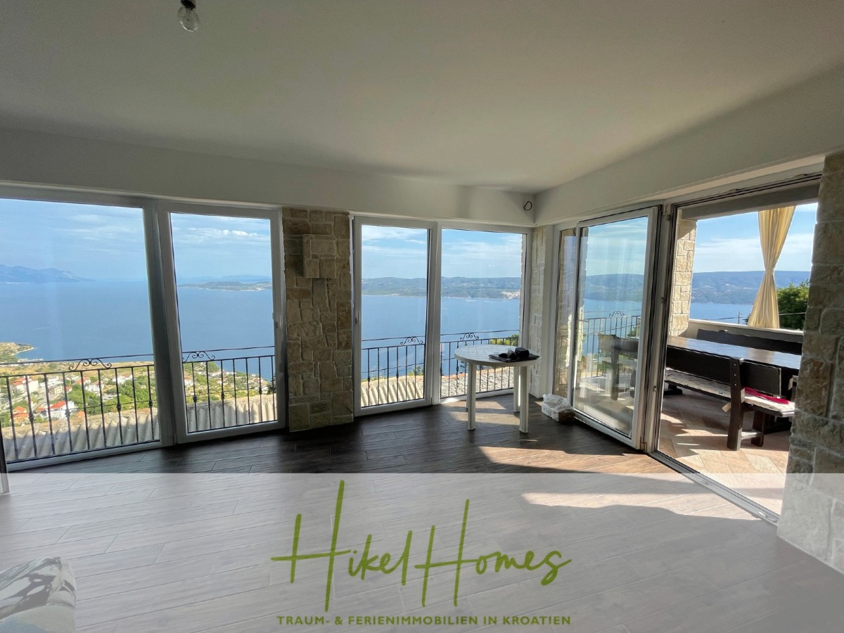 Ein helles Zimmer mit großen Fenstern und Glastüren öffnet sich zu einem Balkon mit Panoramablick auf das Meer und die Küste. Modernes Mobiliar und Steinsäulen sorgen für Stil. Der Schriftzug "Hikel Homes" erscheint am Boden. - Meerblick aus perfekter Höhenlage mit 3 Schlafzimmern