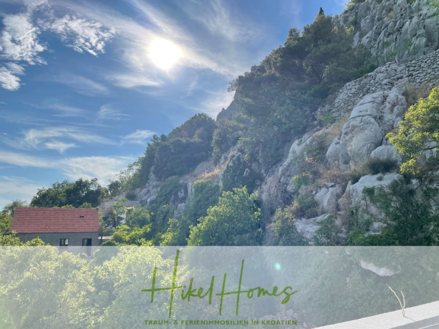 Meerblick aus perfekter Höhenlage mit 3 Schlafzimmern - Ein sonniger Hang in Kroatien mit felsigem Gelände, grünen Bäumen, einem kleinen Haus mit rotem Dach und einem strahlend blauen Himmel mit Schäfchenwolken. Der überlagerte Text lautet: "Hikel Homes - Traum & Ferienimmobilien in Kroatien. (46076)