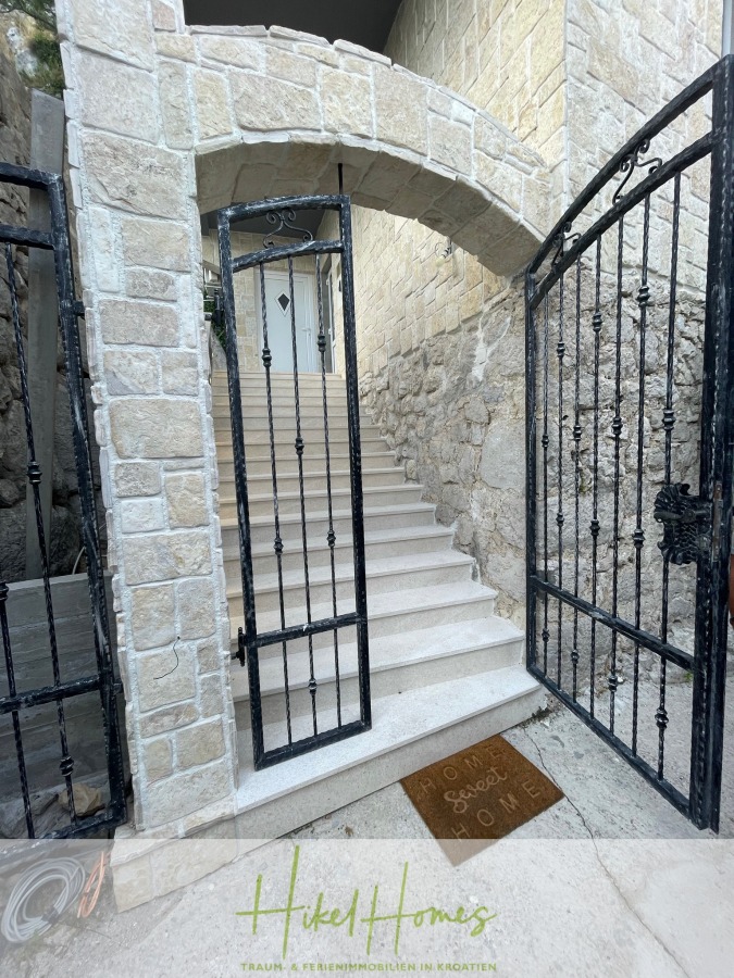 Meerblick aus perfekter Höhenlage mit 3 Schlafzimmern - Ein offenes schmiedeeisernes Tor gibt den Blick auf eine Steintreppe frei, die zu einer weißen Tür führt. Auf einer braunen Fußmatte am Eingang steht "Our Sweet Home". Die Wände und Stufen sind aus hellem Stein. (46088)