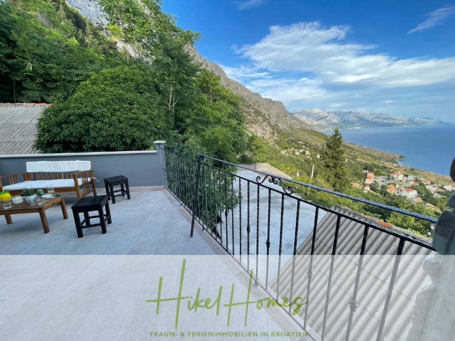 Meerblick aus perfekter Höhenlage mit 3 Schlafzimmern - Eine geräumige Terrasse mit Holzmöbeln und Erfrischungen bietet einen Blick auf üppige Hügel, verstreute Häuser und das blaue Meer unter einem teilweise bewölkten Himmel in Kroatien. Das Bild hat den Text "Hikel Homes" am unteren Rand. (46085)