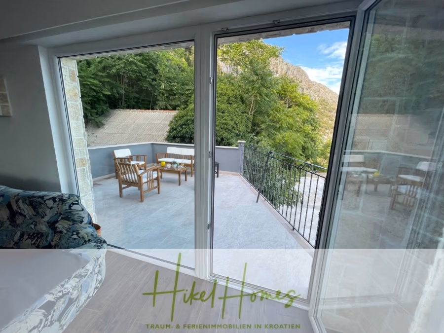 Meerblick aus perfekter Höhenlage mit 3 Schlafzimmern - Geräumige Terrasse mit Holzstühlen und einem Tisch, umgeben von Grün und Hügeln, von einem Zimmer aus durch eine Glastür gesehen. Das Bild enthält den Text „Hikel Homes – Traum- & Ferienimmobilien in Kroatien“. (41486)
