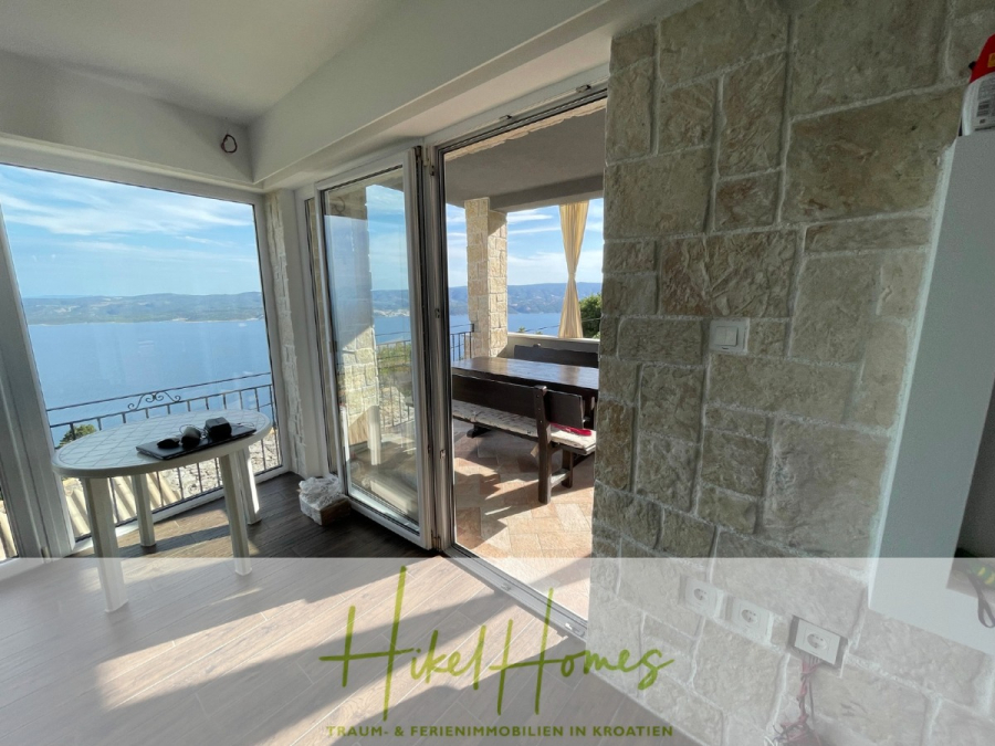 Meerblick aus perfekter Höhenlage mit 3 Schlafzimmern - Ein heller Raum mit großen Glastüren öffnet sich zu einer Steinterrasse mit einer Holzbank und einem atemberaubenden Blick auf das Meer und die fernen Hügel. Sonnenlicht erfüllt das moderne Interieur, und ein kleiner Tisch steht am Fenster. (46078)