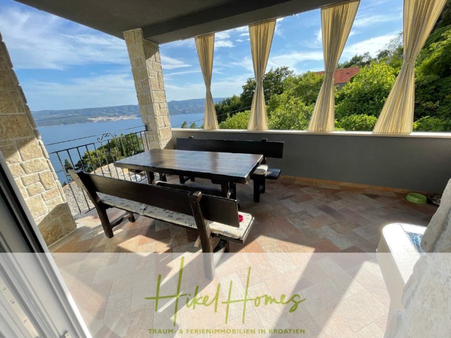 Meerblick aus perfekter Höhenlage mit 3 Schlafzimmern - Eine überdachte Terrasse mit einem Holztisch und Bänken bietet einen Blick auf das Meer und die Hügel. Helle Vorhänge hängen zwischen Steinsäulen, und das Sonnenlicht erhellt den gefliesten Boden. Die Szene erinnert an ein friedliches Ferienhaus in Kroatien. (46080)