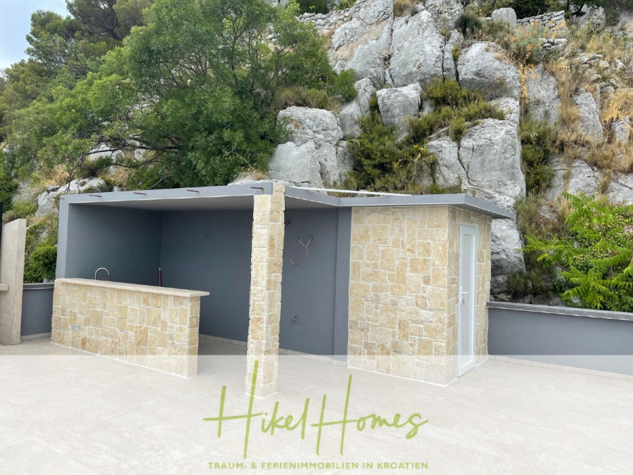 Meerblick aus perfekter Höhenlage mit 3 Schlafzimmern - Eine Außenküche und ein kleines Gebäude mit Steinmauern und einem Flachdach vor einem felsigen, begrünten Hang. Der Text "Hikel Homes" und "Traum- & Ferienimmobilien in Kroatien" erscheint am unteren Rand. (46070)