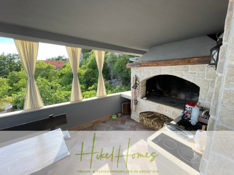 Meerblick aus perfekter Höhenlage mit 3 Schlafzimmern - Überdachte Außenterrasse mit Steingrill, gestapeltem Brennholz, einem Kochfeld und einem Esstisch aus Holz. Der Raum ist zum Grün hin offen und zwischen den Säulen hängen gelbe Vorhänge. (46084)