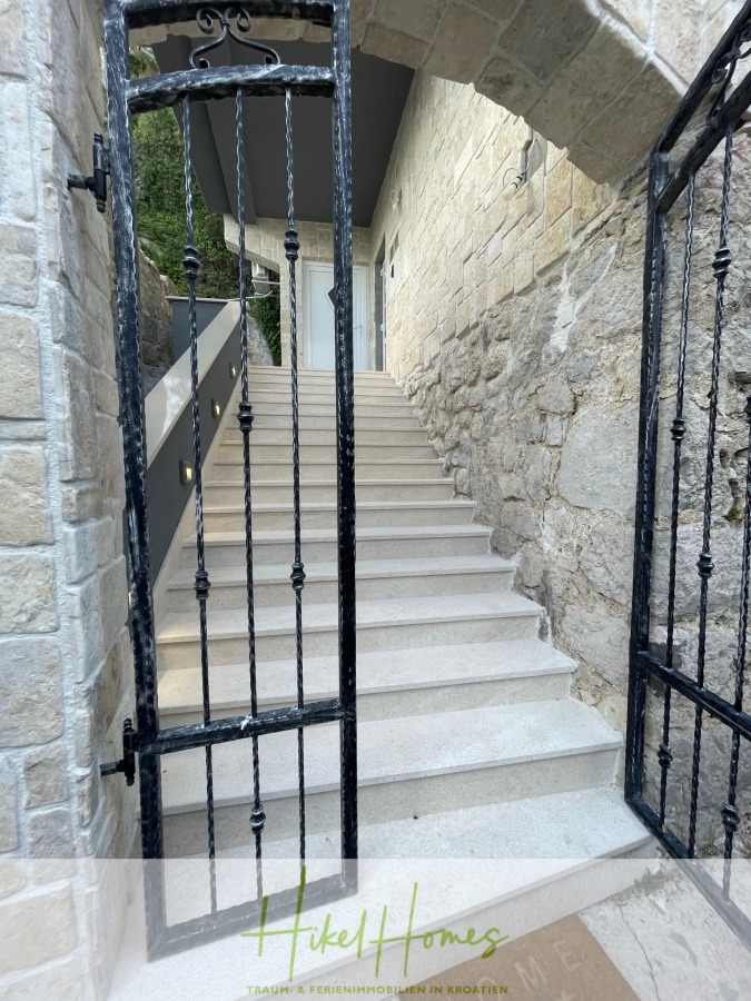 Meerblick aus perfekter Höhenlage mit 3 Schlafzimmern - Ein schwarzes schmiedeeisernes Tor führt zu einer Steintreppe, die zu einem modernen Steingebäude hinaufführt. Die Treppe ist von Steinmauern eingefasst und mit kleinen eingebauten Lampen versehen. Am unteren Ende stehen die Worte "Hikel Homes". (46089)