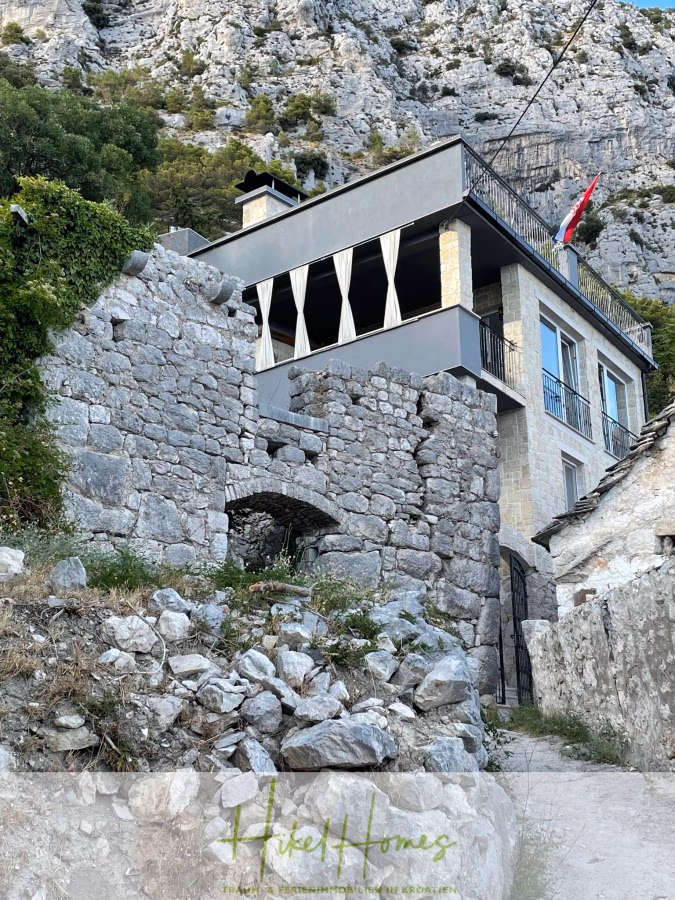 Meerblick aus perfekter Höhenlage mit 3 Schlafzimmern - Ein Steinweg führt zu einem modernen Haus mit großen Fenstern und einer Terrasse, das an einem felsigen Hang erbaut wurde. Alte Steinmauern säumen den Weg. Am Haus ist eine Flagge zu sehen, und zwischen den Felsen verstreut sich Grün. (41463)