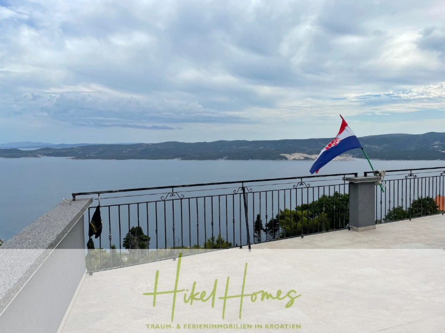 Meerblick aus perfekter Höhenlage mit 3 Schlafzimmern - Eine Terrasse mit einem Metallgeländer bietet einen Blick auf ein ruhiges blaues Meer und ferne Hügel unter einem bewölkten Himmel; eine kroatische Flagge weht auf dem Geländer. Der Text am unteren Rand lautet "Hikel Homes - Traum- & Ferienimmobilien in Kroatien. (46069)